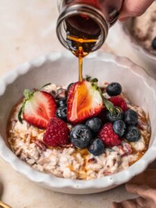 Maple syrup being poured into a bowl of bircher muesli and berries.