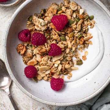 Toasted vanilla almond granola over yoghurt, topped with raspberries in a white bowl.