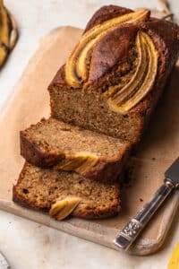 A loaf of gluten free dairy free banana bread on a wooden board, with a fresh banana off to the side.