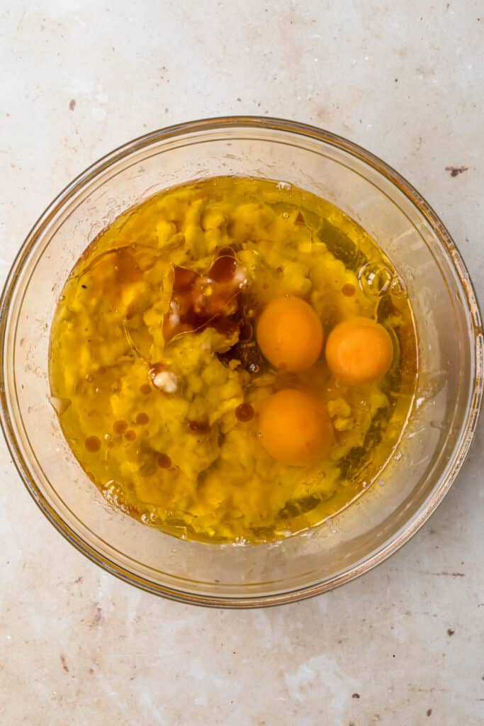 Eggs, olive oil and vanilla extract added to mashed banana in a glass mixing bowl.