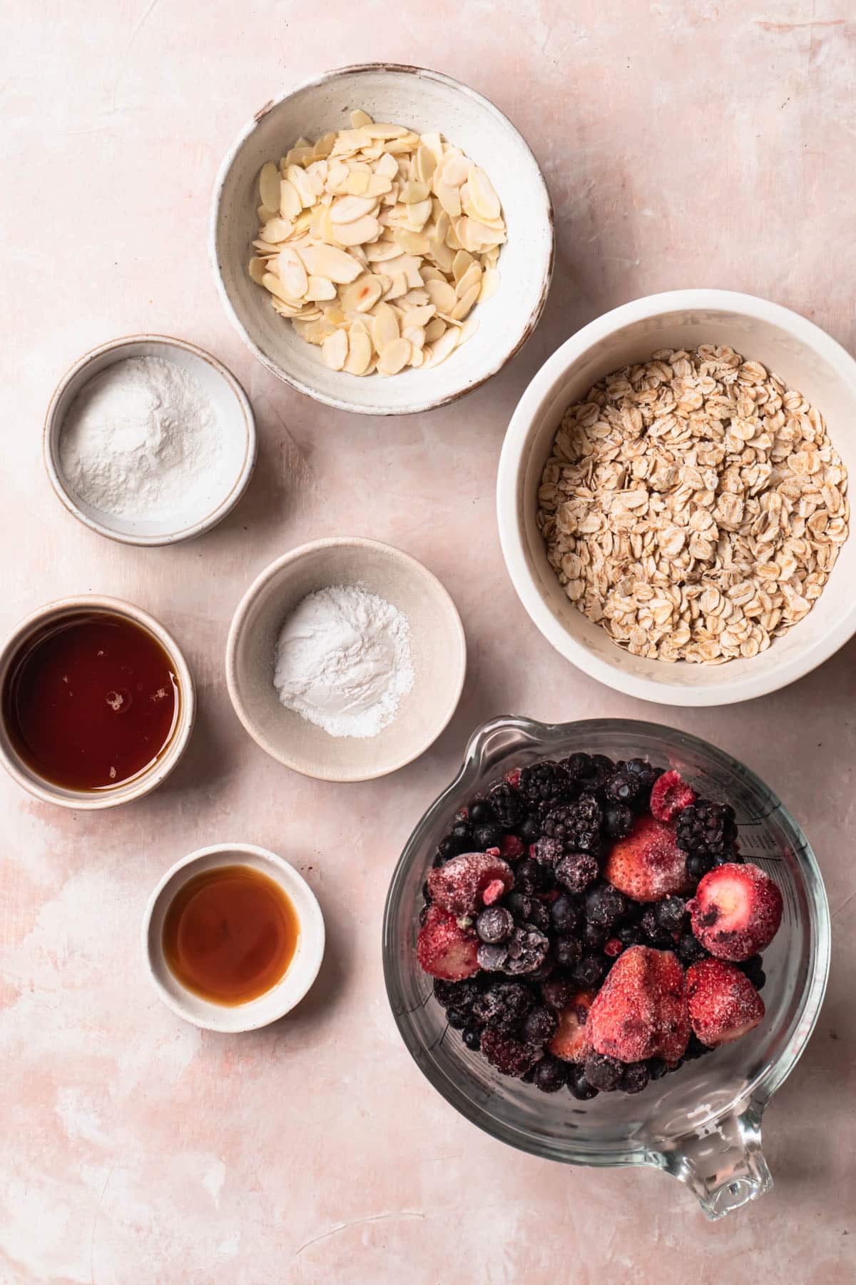 Ingredients for berry breakfast crumble laid out in individual bowls on a pink backdrop.