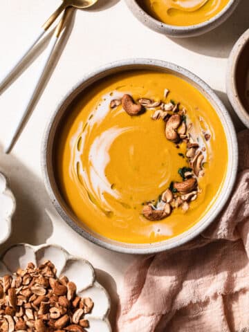 spicy Thai pumpkin soup in a bowl topped with coconut milk and toasted cashews, on a peach background.