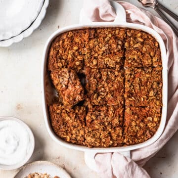 Carrot cake baked oats in a white baking dish, with a small bowl of coconut yoghurt and walnuts.