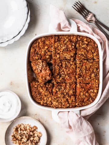Carrot cake baked oats in a white baking dish, with a small bowl of coconut yoghurt and walnuts.