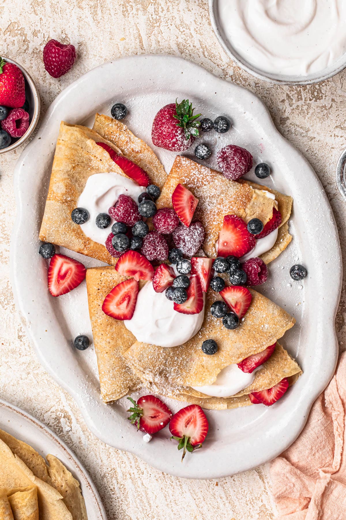 Almond flour crepes topped with fresh berries, coconut yoghurt and icing sugar on a large platter.