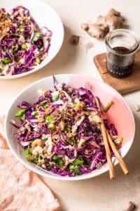 Asian coleslaw recipe served in two pink and white bowl, with chopsticks resting on the bowl at the front of the image.