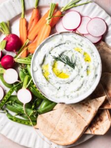 paleo tzatziki on a platter with flat bread, baby carrots, radish and sweet peas, pictured on a pink background.
