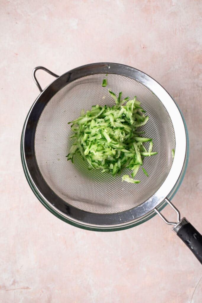 grated cucumber in a sieve sitting over the top of a glass bowl