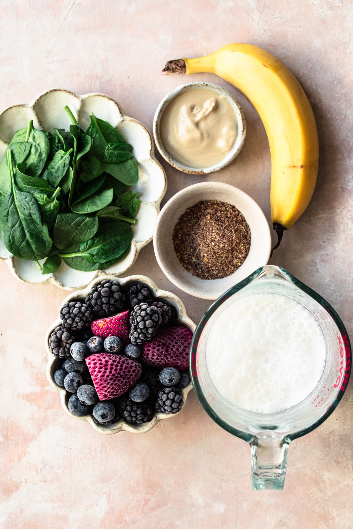 Ingredients for spinach berry smoothie including spinach, berries, coconut milk, flaxseed meal and tahini, laid out in individual bowls on a pink backdrop.