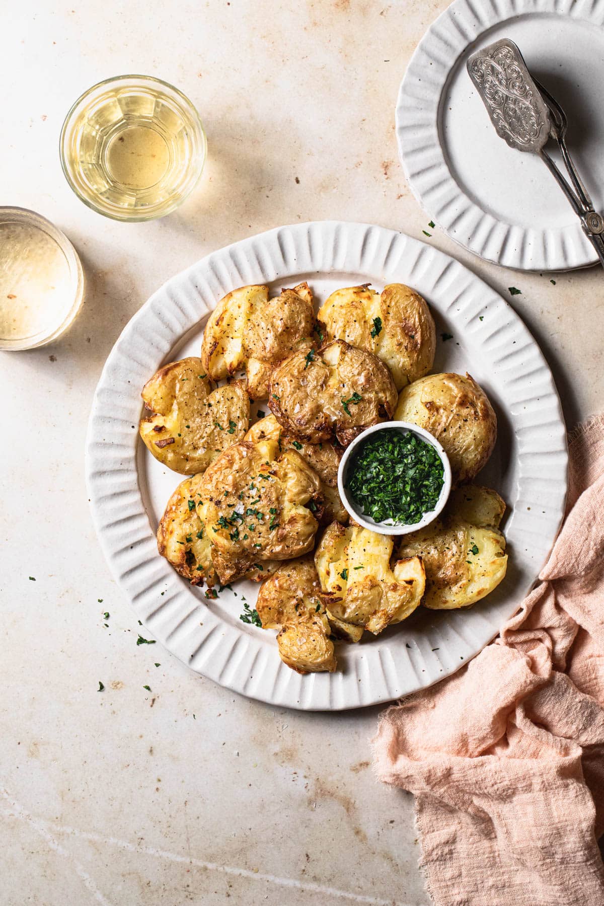 air fryer smashed potatoes on decorative white plate, with small bowl of chopped herbs and two glasses of white wine.