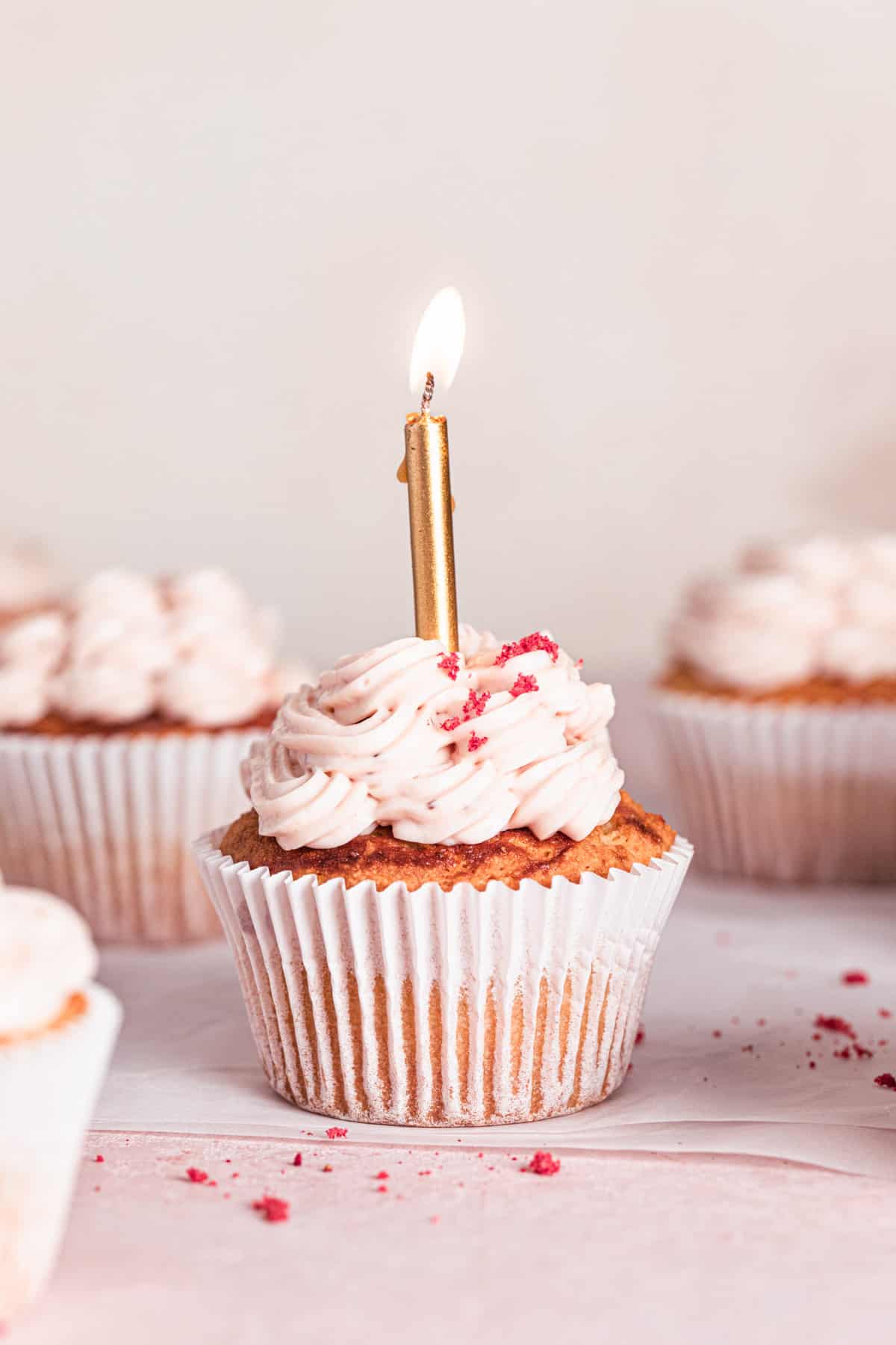 gluten free strawberry cupcakes on a light peach and pink backdrop with a lit birthday candle in the cupcake