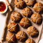 Gluten free hot cross buns on a baking paper lined baking tray, a small bowl of glaze to the side of the tray as well as a basting brush.
