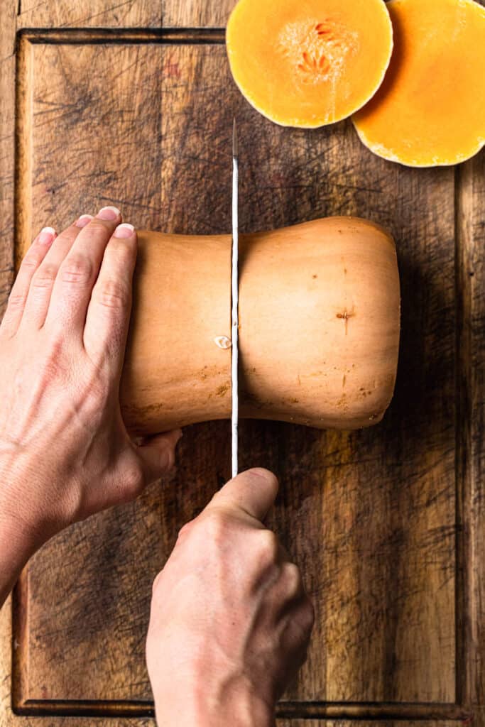 Butternut pumpkin on a timber chopping board, ends removed, being sliced in half.