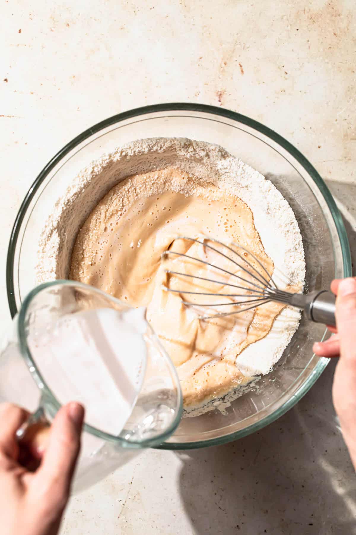 Pouring milk into flour to make pancake batter mixture while whisking.