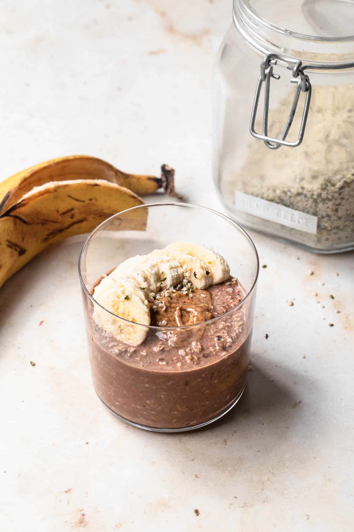 Peanut butter chocolate overnight oats in a glass tumbler, topped with banana slices and hemp seeds.
