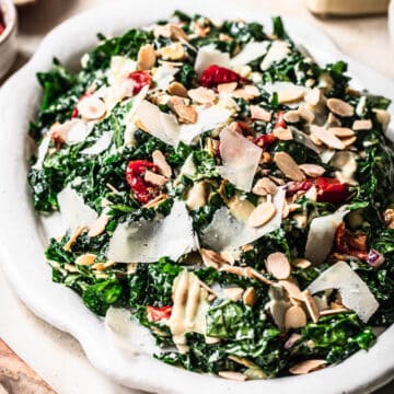Kale salad with shaved parmesan cheese, semi dried tomatoes, toasted almonds and tahini dressing on a white serving platter.