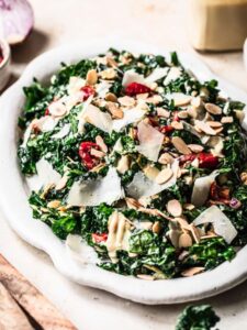 Kale salad with shaved parmesan cheese, semi dried tomatoes, toasted almonds and tahini dressing on a white serving platter.