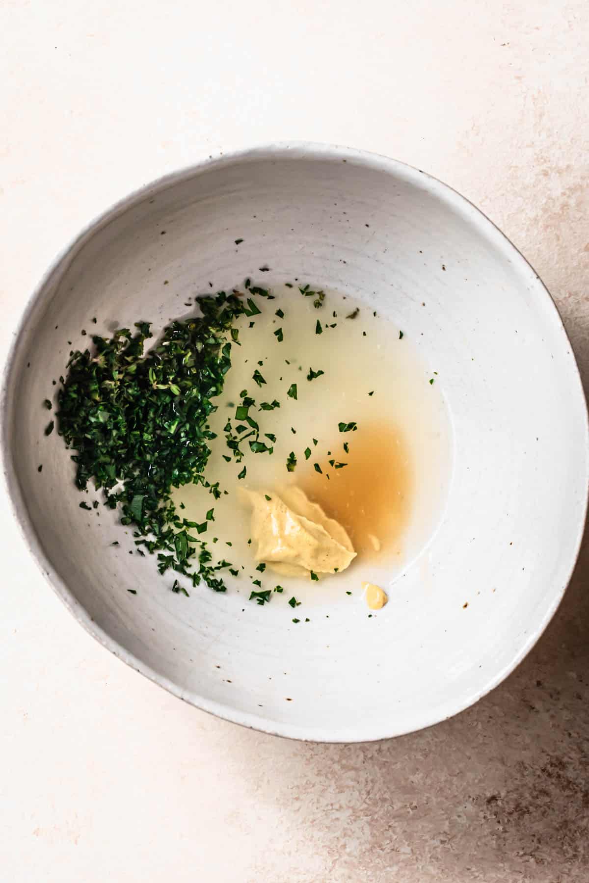 Top down image of ingredients for a lemon herb dressing in a white ceramic bowl.