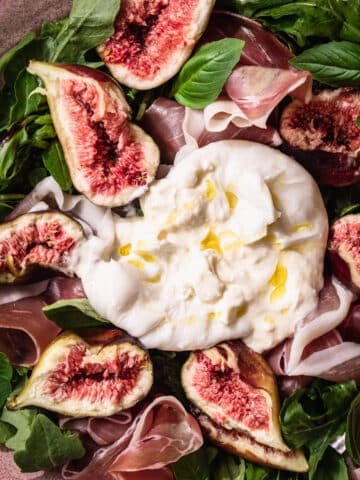 Close up image of fig, prosciutto, rocket and burrata salad. The burrata ball is in the centre and sliced open so that the creamy centre oozes out.