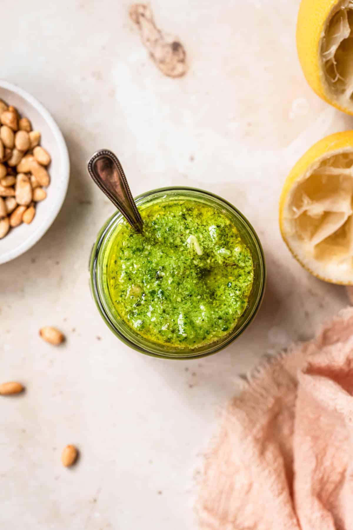 Top down photo of pesto salad dressing in a small jar with squeezed lemons and pine nuts in the background.