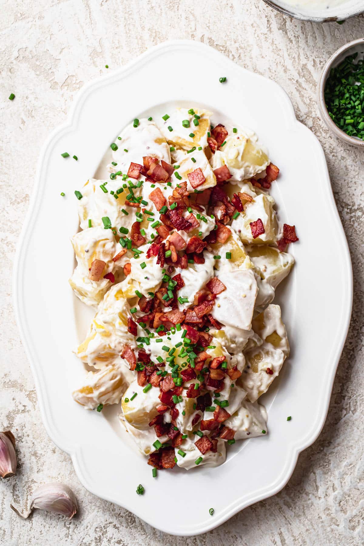 Creamy potato salad served on a white platter, topped with crispy bacon bits and finely sliced chives.