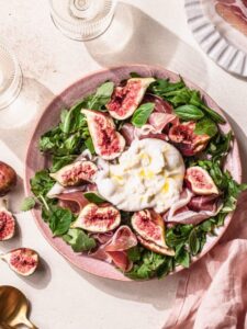 Fig and burrata salad with prosciutto in a pink bowl with two glasses of wine in the background.