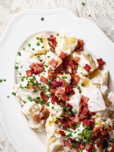Creamy potato salad served on a white platter, topped with crispy bacon bits and finely sliced chives.
