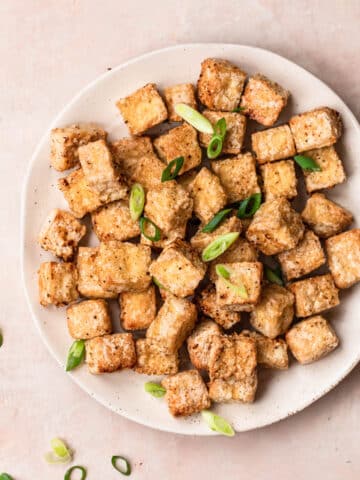 Golden brown salt and pepper tofu cooked in the air fryer, on a white plate, topped with thinly sliced green onions.