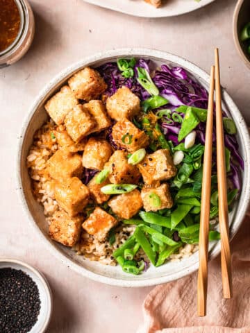 Crispy tofu bowl with thinly sliced snow peas and cabbage, topped with thinly sliced green onions. The bowl has chopsticks placed on top, with sauce and crispy tofu to the side.