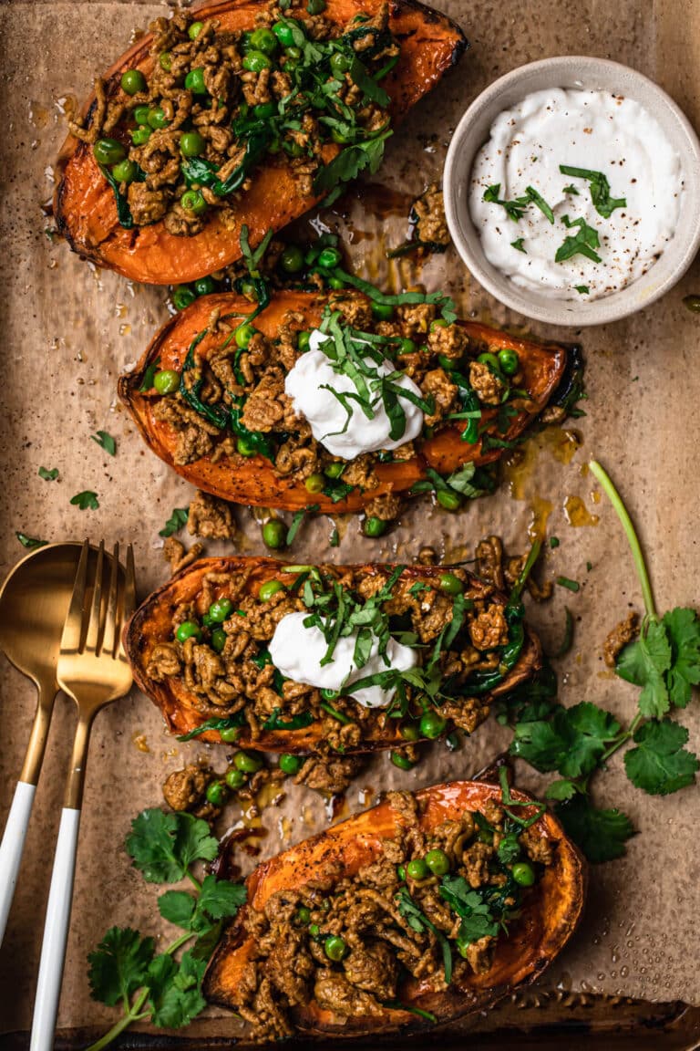 Moroccan lamb mince stuffed sweet potatoes on a brown parchment paper lined baking tray. The stuffed sweet potatoes are topped with yoghurt and thinly sliced fresh cilantro.