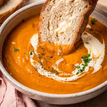 A slice of buttered bread being dipped into a bowl of sweet potato pumpkin soup.