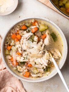 A bowl of chicken noodle soup, with a spoon resting in the bowl, a small bowl of shaved parmesan to the side.