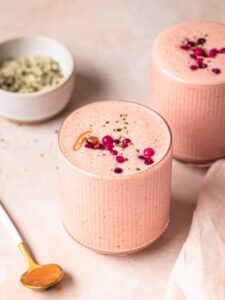 Fresh cranberry smoothie in a ribbed glass tumbler, with a second smoothie and a small bowl of hemp seeds in the background.