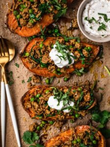 Moroccan lamb mince stuffed sweet potatoes on a brown parchment paper lined baking tray. The stuffed sweet potatoes are topped with yoghurt and thinly sliced fresh cilantro.