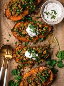 Moroccan lamb mince stuffed sweet potatoes on a brown parchment paper lined baking tray. The stuffed sweet potatoes are topped with yoghurt and thinly sliced fresh cilantro.