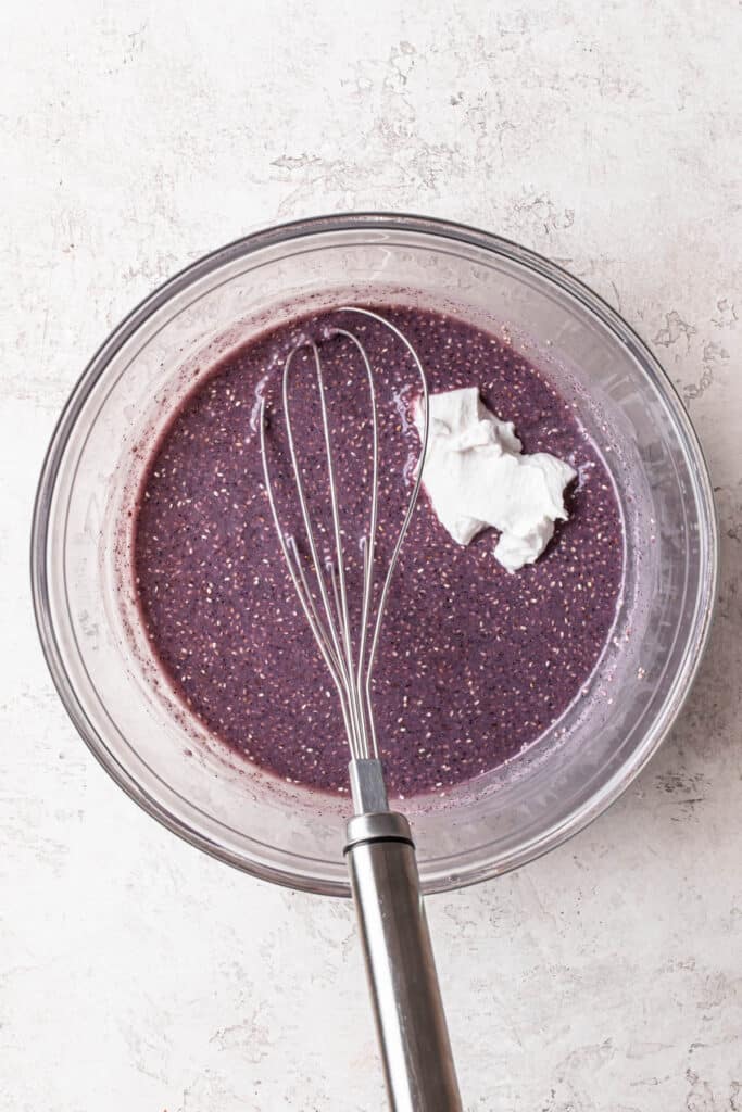 Yogurt added to a glass mixing bowl filled with blueberry chia pudding.
