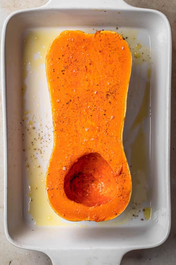 Halved butternut squash with seeds removed, drizzled with olive oil and seasoned with salt and pepper, in a large white baking dish.