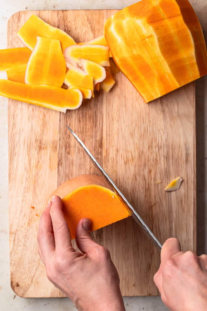 Image of a hand holding a piece of butternut squash, standing up on a chopping board, using a sharp knife in the other hand to slice the skin off.