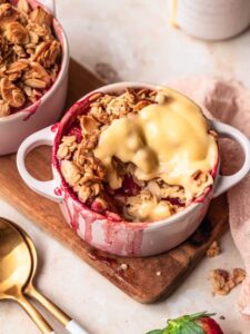 Strawberry apple crumble in a small ramekin, topped with custard with a scoop taken out of the crumble.