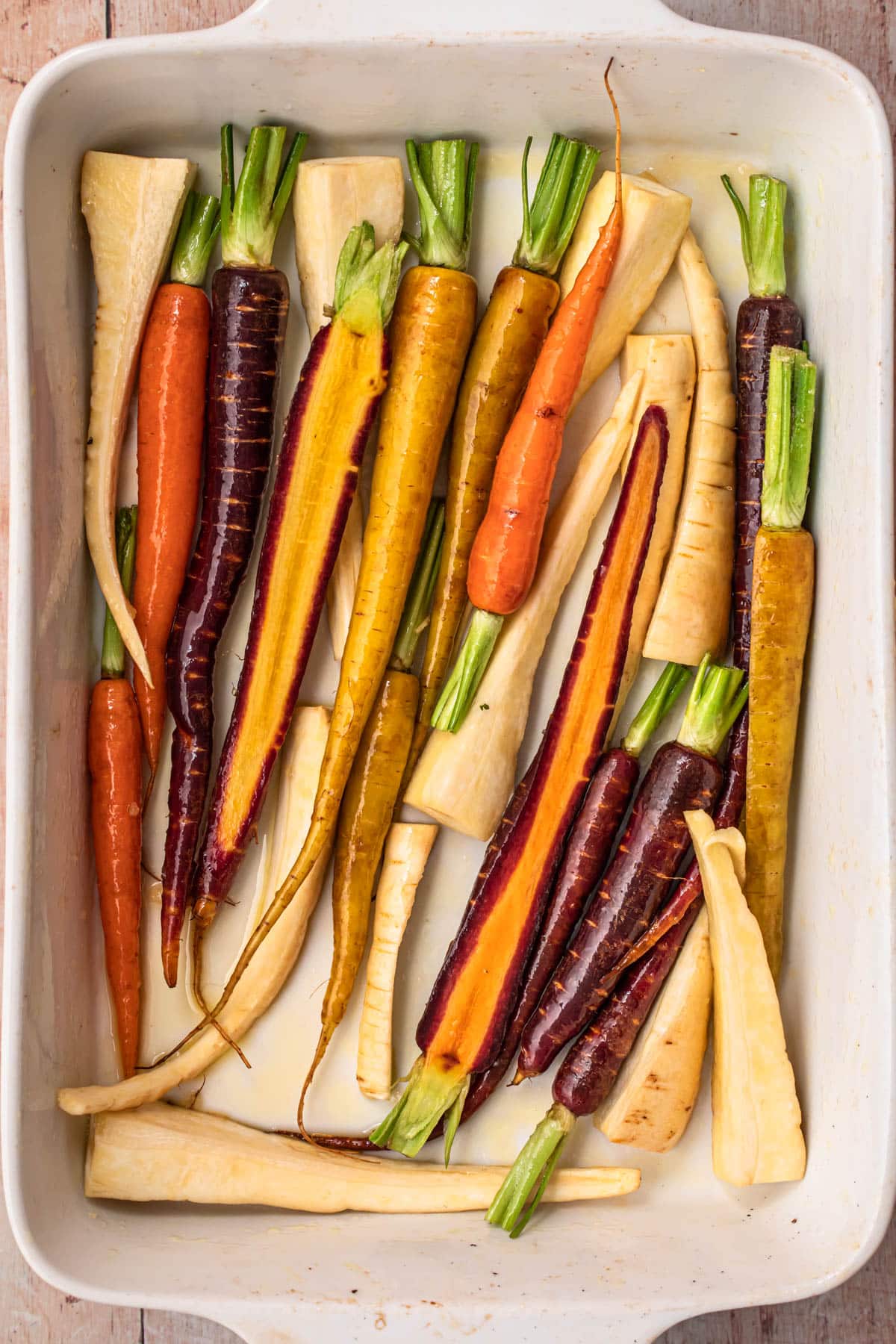 Baby carrots and sliced parsnips spread out in a single layer in a large white baking dish.