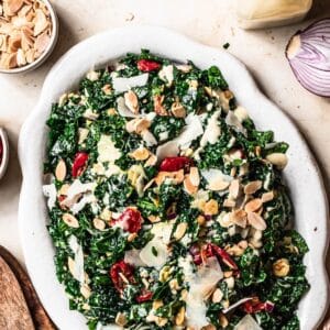 Kale salad with shaved parmesan cheese, semi dried tomatoes, toasted almonds and tahini dressing on a white serving platter.
