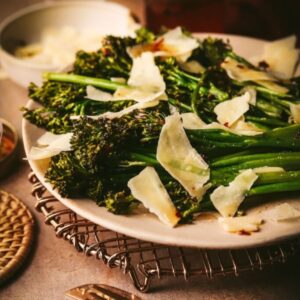 Long stemmed broccoli on a plate on a set table with cheese shavings and oil olive on top.