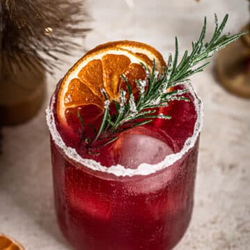 Festive Cranberry Rosemary cocktail in a glass with ice, topped with dehydrated orange slices and rosemary.