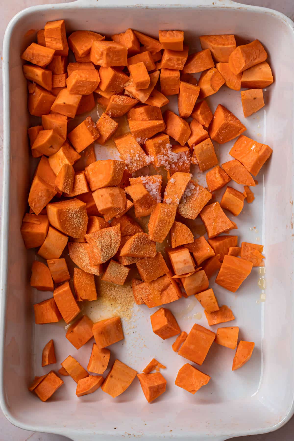 Sweet potato cubes in a large white baking dish with olive oil and seasoning.