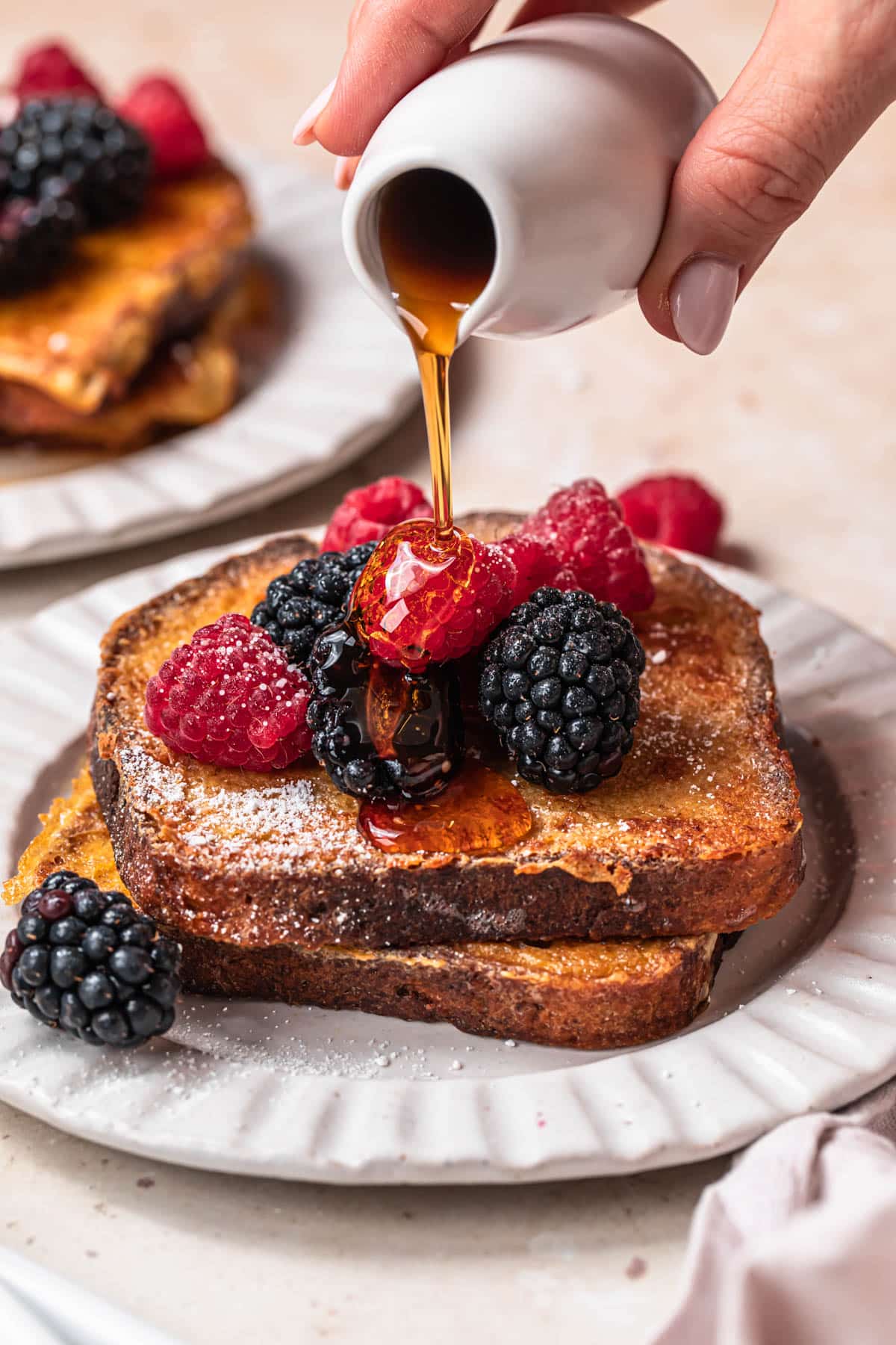 Almond milk French toast on a plate, topped with berries with maple syrup being poured over the top.