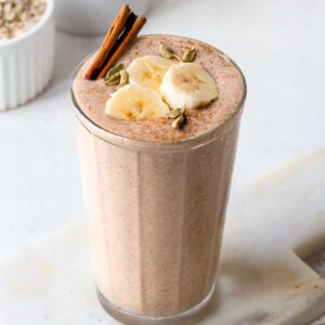 Chai banana smoothie in a ribbed glass topped with sliced bananas, cardamom pods and a cinnamon stick.