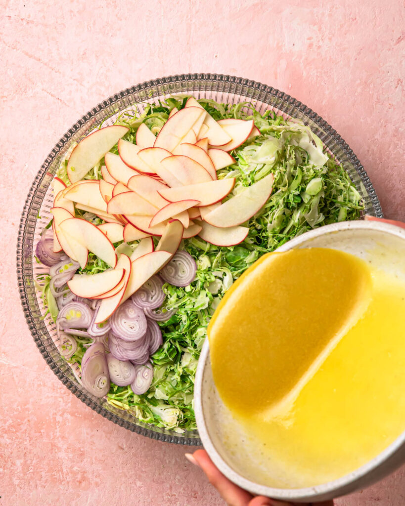 Maple dijon dressing being poured into a large bowl with shaved brussels sprouts and thinly sliced apples. 