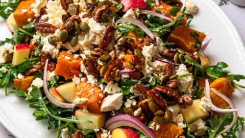 Butternut squash and feta salad topped with feta and candied pecans on a serving platter with dressing in a jug in the background.