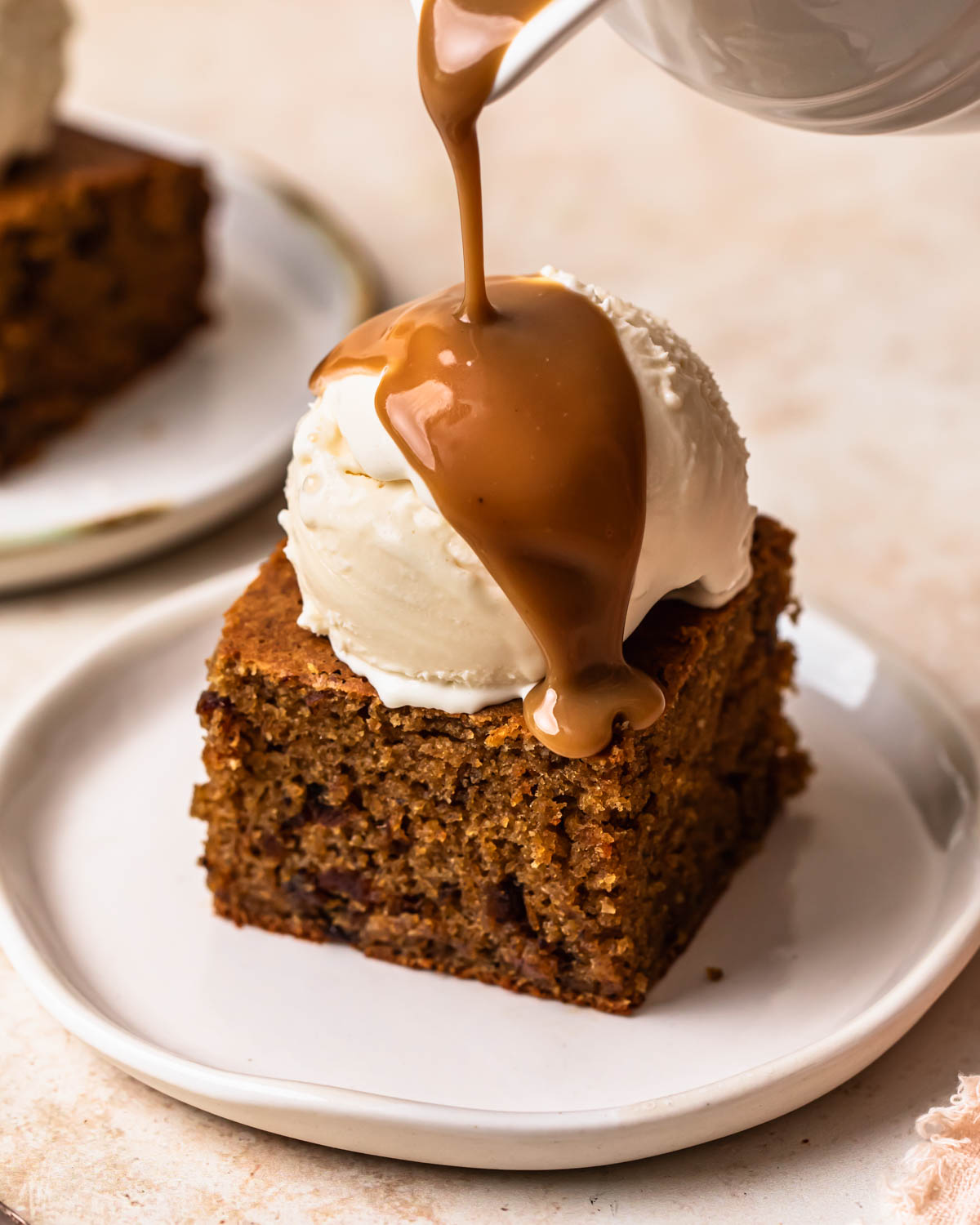 A slice of gluten free sticky date pudding topped with ice cream and warm toffee sauce being poured over the top.