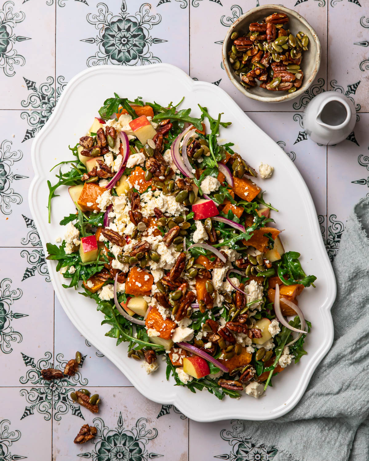 Butternut squash and feta salad topped with feta and candied pecans on a serving platter with dressing in a jug in the background.
